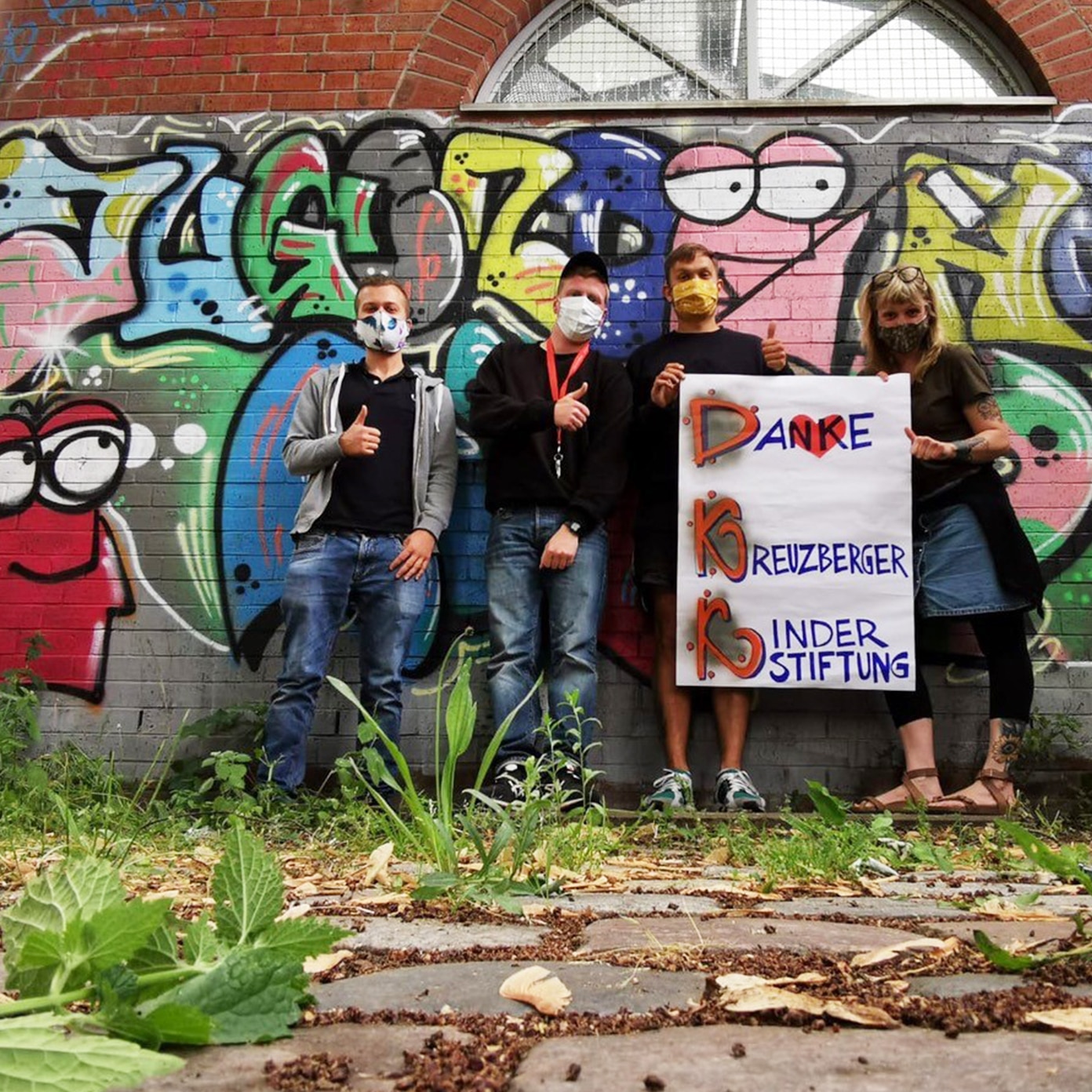 Jugendliche vor Graffiti-Wand mit Mund-Nasen-Schutz und Plakat "Danke Kreuzberger Kinderstiftung"